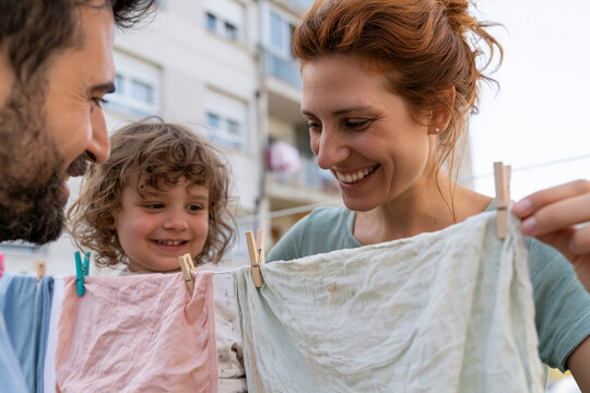 Warm family smiles while hanging laundry outdoors. Child's curls bounce as they watch parents' joy. Soft daylight glows on pink and white clothes
