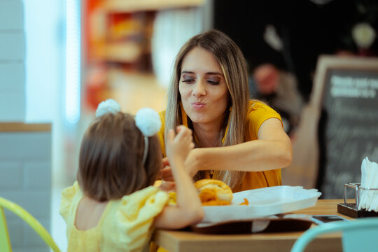 Happy Mother Making Funny Face While Feeding Little Daughter In Cafe. Loving playful mom enjoys sweet dessert together with her cute toddler inside restaurant
