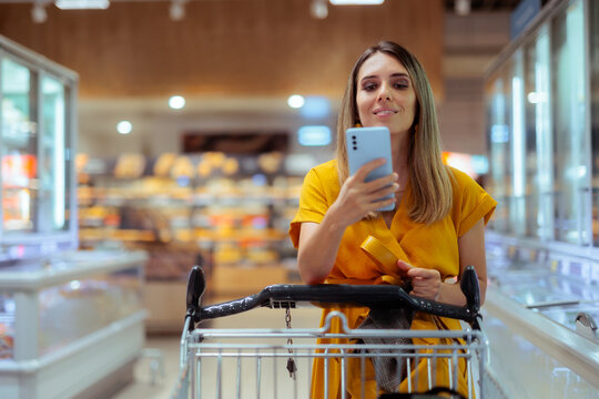 Happy Young Woman Checking Digital Shopping List On Her Smartphone. Smiling female customer looks at mobile phone while holding cart inside modern supermarket