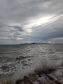 An eye-level wide perspective showing sea waves breaking with white foam against a rocky shore, with a distant island silhouetted on the horizon under a cloudy, overcast sky.