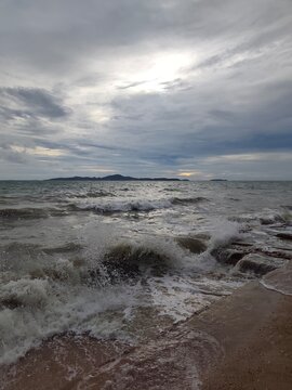 An eye-level wide perspective showing sea waves breaking with white foam against a rocky shore, with a distant island silhouetted on the horizon under a cloudy, overcast sky.