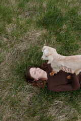 Obraz premium Goat kid resting on a smiling woman lying in grass, outdoors lifestyle moment, playful affection, sunlit meadow scene, natural colors, relaxed pose, gentle bond, carefree mood, rustic charm.