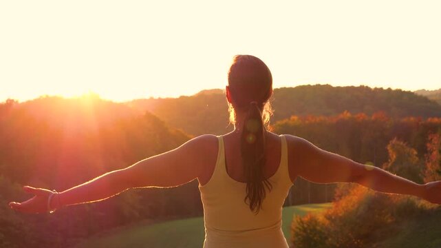 CLOSE UP, LENS FLARE: Woman stands on scenic hillside when she raises her arms toward autumn setting sun. Colourful forested hills surround outdoor moment of celebration and connection with nature.