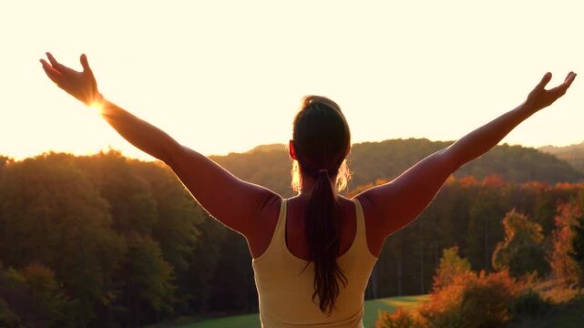 CLOSE UP, LENS FLARE: Woman standing on scenic hill with arms raised toward glowing sunset. She celebrates freedom and connection with nature as golden light spills over autumn woods and rolling hills