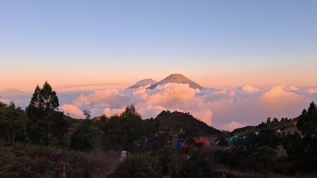 Hikers walking through basecamp overlooking Mount Sindoro at sunrise with clouds covering valley in Central Java Indonesia.