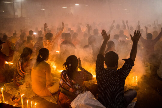 Narayanganj, Dhaka Division, Bangladesh - 09 November 2024: View of a crowd sitting on the ground with hands raised during a candlelit religious ceremony filled with smoke.