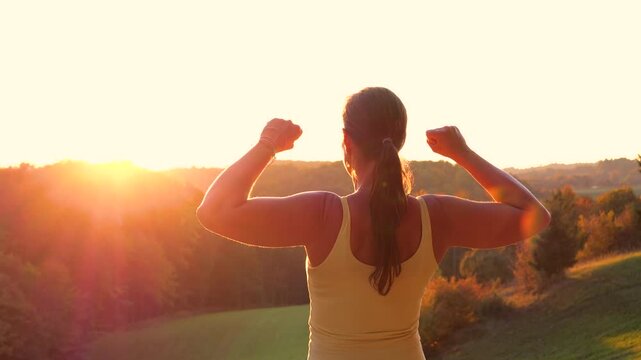 CLOSE UP, LENS FLARE: Back view of young woman raising clenched fists in victorious pose above colourful autumn hills. Powerful moment of achievement and celebration after an active day in nature.