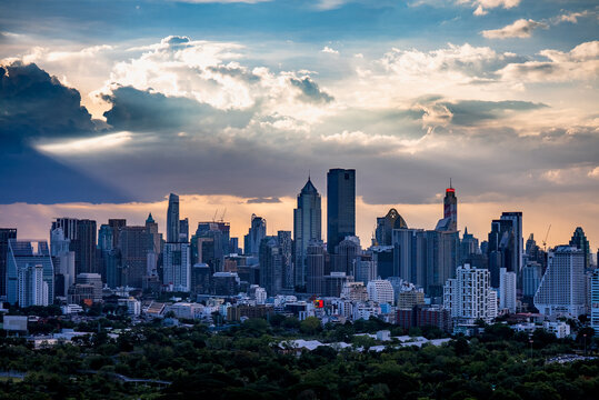 Bangkok, Thailand - 20 June 2024: View of the modern skyline with towering skyscrapers, lush greenery in the foreground, and dramatic sunbeams breaking through clouds at sunset.