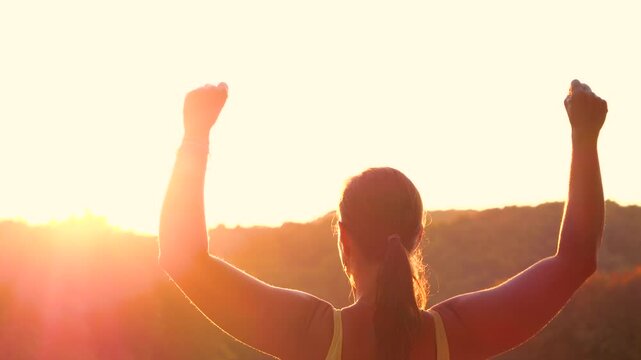 CLOSE UP, LENS FLARE: Young woman lifts her arms in celebration while overlooking glowing autumn countryside. Pristine moment of victory, strength and accomplishment after an active day in nature.