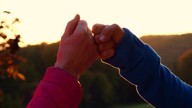 CLOSE UP, LENS FLARE: Two adult people greet each other with a fist bump in warm golden sunlight. Moment of bonding and celebration with forested hills in the background and glowing morning atmosphere