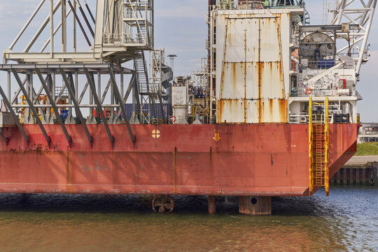 Esbjerg, Denmark - 29 July 2026: View of the Ziton Wind Enterprise crane ship jacked up in the harbor, with its thruster visible under the red hull and industrial structures rising above.
