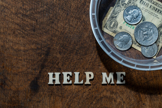 Donation jar with US money and help me text, closeup. Transparent plastic donation box with one dollar bill and coins next to the word help me made of wooden letters on rustic table surface