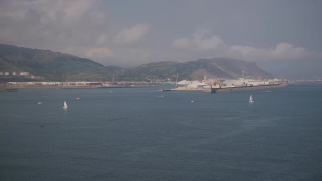 Coastal landscape with sailboats enjoying the calm bay waters, featuring industrial port infrastructure and distant hills