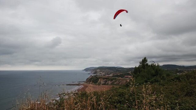 Person paragliding above the scenic coastal landscape with cliffs, beaches, and the Cantabrian Sea under an overcast sky