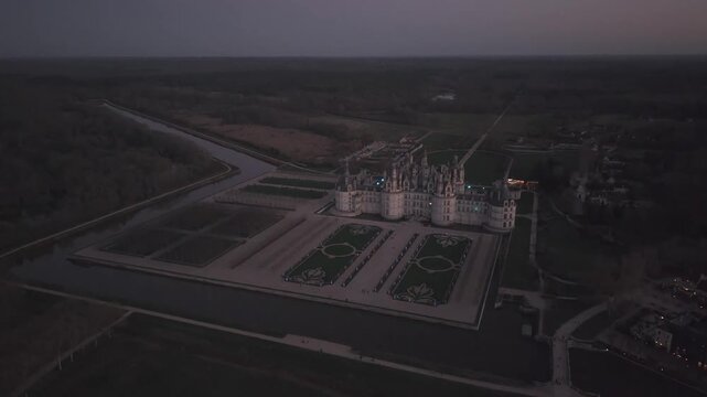 An aerial view at dusk shows Chateau de Chenonceau and its geometric formal gardens glowing softly in the fading light.