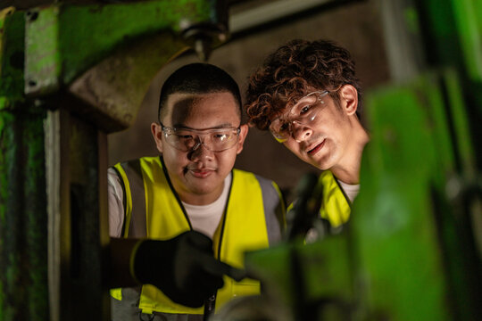 Thai father with curly hair and his teen son inside an old family machine shop, son points a gloved index finger down at the machine mechanism in, family enterprise mentorship