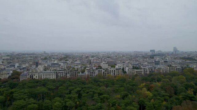 Sweeping aerial drone footage of Paris cityscape unfolds above lush urban park in soft daylight, revealing dense architectural patterns, autumn trees, and serene overcast atmosphere