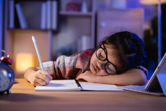 Young girl wearing glasses writing in notebook while resting head on desk at night with tablet device and clock in background cozy study environment