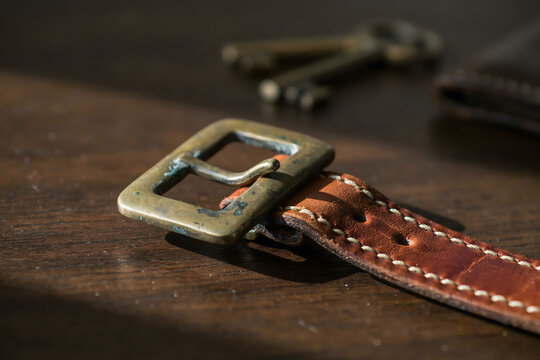 Brown leather watch strap with a brass buckle and vintage keys resting on a dark wooden table surface under warm natural side lighting