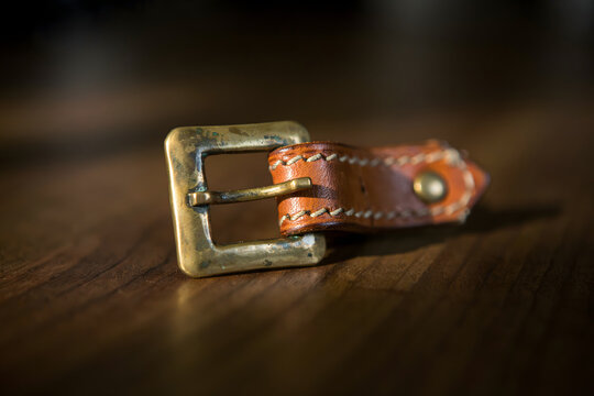 Close up of a brown leather watch strap with a brass buckle resting on a dark wooden surface with warm natural light hitting the