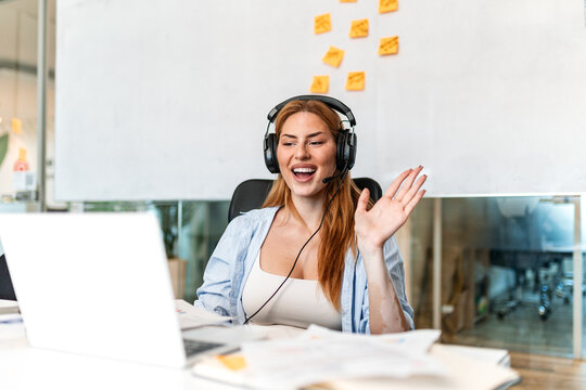 Video conference. Remote communication, Woman with headset waving hand at laptop, Corporate meeting concept. Smiling female employee talking on online call in modern office workspace.