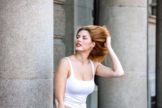 Urban portrait. City fashion style, Young woman touching her hair outdoors, Modern lifestyle and beauty concept. Attractive redhead female posing near stone architecture in city center.