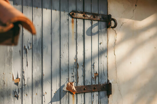 Rustic wooden window shutters painted light blue with visible peeling texture and heavy metal hinges attached to a weathered white stucco wall