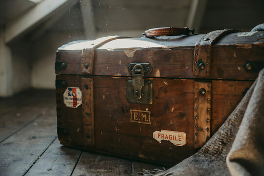Vintage wooden travel chest with metal reinforcements and paper labels sitting on a dark wooden floor in a dimly lit attic environment