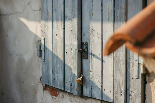 Weathered light blue wooden window shutters with a central metal latch and a terracotta roof tile corner visible against a white stucco wall