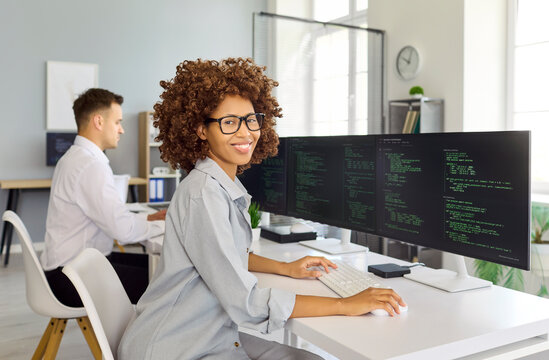 Woman programmer developer coding at computer in office. Bright workspace with ultrawide screens showing code and a coworker nearby supporting teamwork. Modern tech workplace concept today.