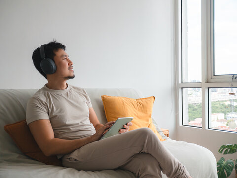 Content Asian man enjoying music with headphones and tablet while relaxing on a comfortable sofa.