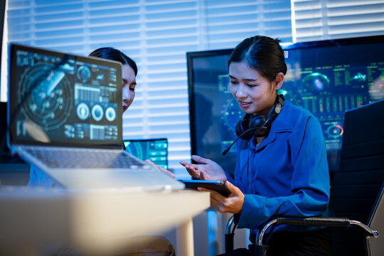 Two women working with digital screens and a tablet in a modern office, discussing data and monitoring systems in a blue lit technology room