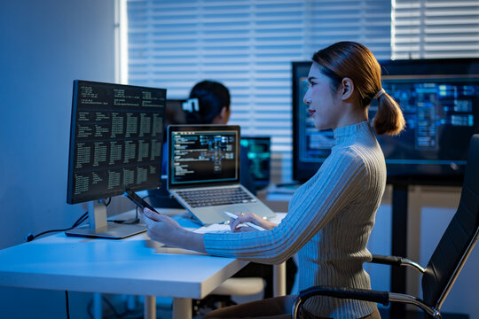 Woman working at a desk with multiple monitors and a laptop in a modern office, focused on coding or data analysis in blue light.