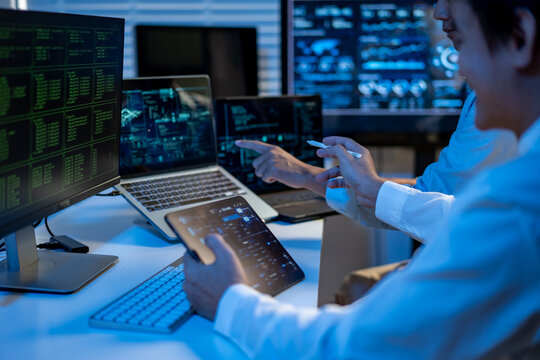 Cybersecurity specialist monitoring data on multiple screens in a dark office, using laptop and tablet to analyze code and network activity.