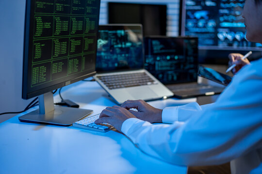 Person working at a desk with multiple monitors showing code and data, typing on a keyboard in a modern office with blue light