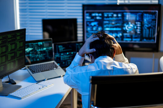 Stressed office worker holding his head while facing multiple computer screens in a modern control room, showing work pressure, data overload and technology problems.