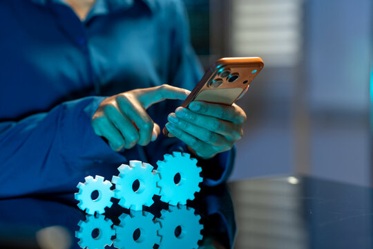Close up of hands using a smart phone beside white gears on a desk, showing digital business automation and online work