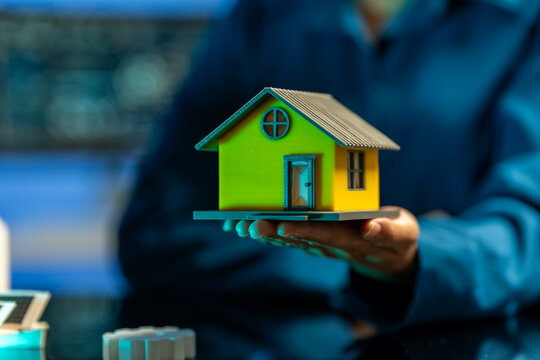Hand holding a small model house with blurred person in the background, symbolizing home buying, property, and real estate investment