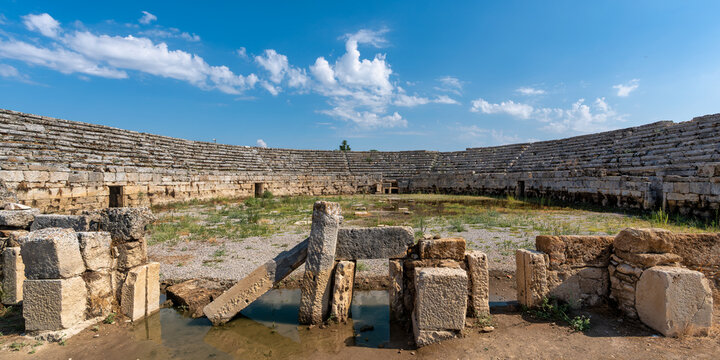 Ancient stone stadium ruins at Perge, a historic Roman archaeological site in Antalya, Turkey, featuring tiered seating and an open arena area under a sunny blue sky