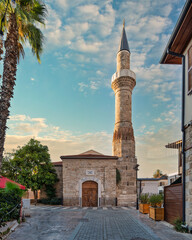 Fototapeta premium The historic Kesik Minare Mosque with its prominent minaret in the old town of Kaleici, Antalya, Turkey, showcasing stone architecture, palm trees, and a cobblestone street at sunset