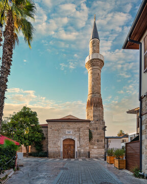 The historic Kesik Minare Mosque with its prominent minaret in the old town of Kaleici, Antalya, Turkey, showcasing stone architecture, palm trees, and a cobblestone street at sunset