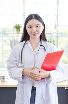 Asian confident young woman doctor wearing medical coat stethoscope standing holding clipboard in modern healthcare office professional physician services hospital clinic wellness trust patient care.