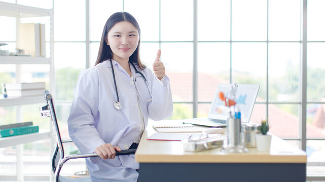 Asian smiling young woman doctor wearing white coat stethoscope giving thumbs up medical success working laptop in healthcare office professional physician services hospital clinic wellness patient.