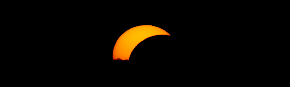 Partial solar eclipse above a dark mountain horizon, with the Moon covering part of the orange Sun as the remaining solar crescent appears to set behind the landscape.
