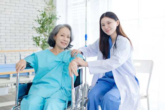 Asian female doctor consulting senior woman patient sitting in wheelchair in hospital clinic attentive comfort support quality elderly healthcare nursing assistance recovery wellness medical services.