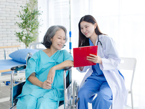 Asian female doctor consulting senior woman patient sitting in wheelchair in hospital clinic attentive comfort support quality elderly healthcare nursing assistance recovery wellness medical services.