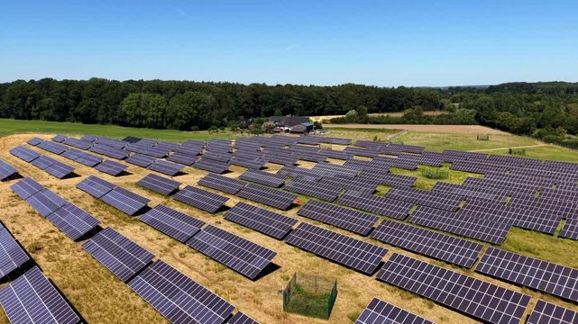 Rows of solar panels stretch across golden farmland in rural United States, dense forest lines background as farmhouse buildings and green fields border wide panel array. Aerial drone approaching shot