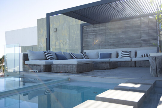Wicker L-shaped sectional sofa is sitting under metal pergola casting linear shadows, beside pool