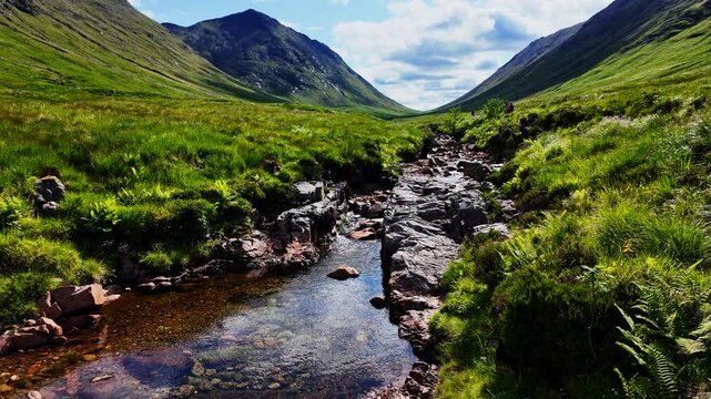 Beautiful close up of a calm stream in the scenic Glencoe Highlands of Scotland on a sunny, summer day. Hikers are walking over a nearby trail in the gorgeous green valley