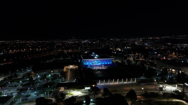 High-angle panoramic view of the Knesset building illuminated with vibrant blue lights against the dark urban backdrop of Jerusalem at night.